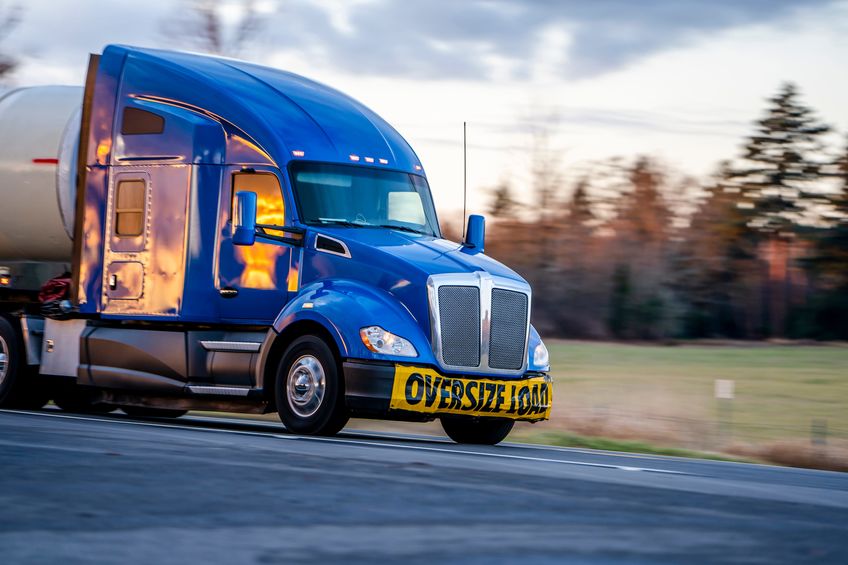 big rig blue bonnet long haul industrial professional semi truck with sign oversize load on the front bumper transporting cargo in long tank semi trailer driving on the road with sunset